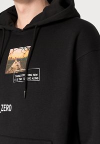 Czarna bluza z kapturem z centralnym nadrukiem przedstawiającym krajobraz i dwoma polami tekstowymi. Materiał wydaje się miękki, z ozdobami w postaci sznureczków przy kapturze.