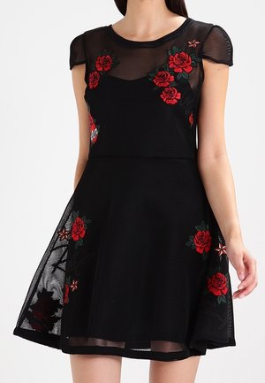 Czarna sukienka z siateczki z haftem czerwonej róży i gwiazdy, krótkie rękawy, dopasowany gorset i rozkloszowana spódnica, noszona przez stojącą osobę.