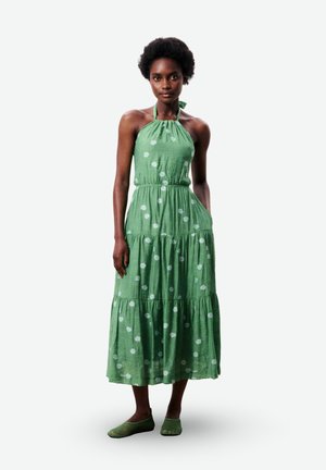 Femme aux cheveux courts bouclés portant une robe verte sans manches à col halter avec un imprimé floral blanc et des ballerines vertes en mesh, debout avec une main dans la poche.