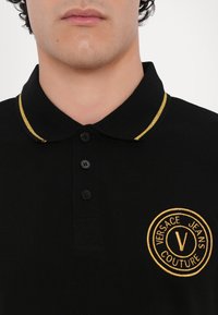 Polo negru dintr-un material texturat, având un logo brodat în aur și cusături de accent pe guler. Patru nasturi negri.