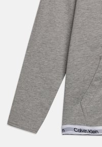Grey sweatshirt with a soft texture, long sleeves, and a pocket. Features a white band at the bottom with "Calvin Klein" in black lettering.