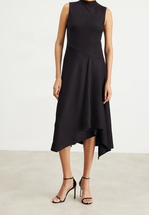 Robe midi noire sans manches avec un ourlet asymétrique portée avec des sandales à talons hauts noires à bride cheville sur un sol clair et un fond uni.