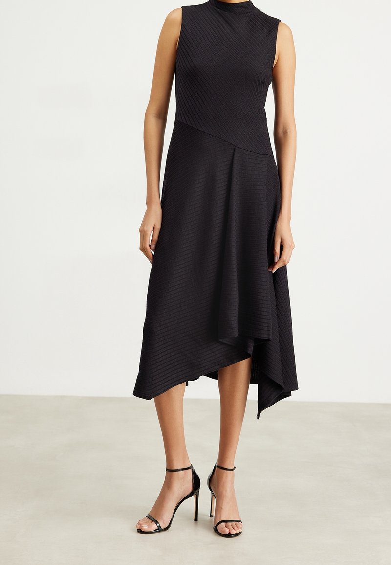 Robe midi noire sans manches avec un ourlet asymétrique portée avec des sandales à talons hauts noires à bride cheville sur un sol clair et un fond uni.