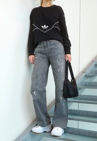 Person wearing black Adidas sweatshirt, gray paint-splattered wide-leg jeans, white platform sneakers, holding black handbag, standing on stairs.