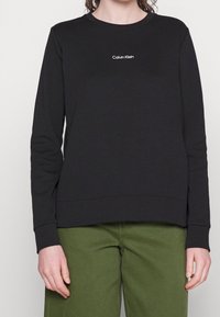 Persona che indossa una maglia nera a maniche lunghe con il logo "Calvin Klein" e pantaloni verdi, in piedi davanti a uno sfondo semplice.