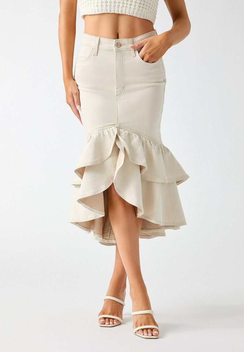 Femme portant une jupe beige taille haute avec un ourlet asymétrique à volants et des sandales blanches à talons avec brides, debout devant un fond uni.