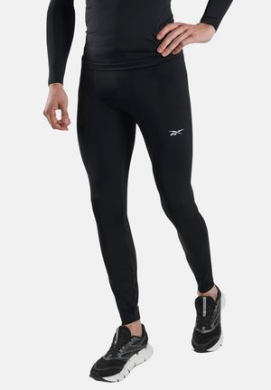 Mann in schwarzen Sportleggings und schwarzen Laufschuhen mit weißen Sohlen, steht mit einer Hand in der Hüfte vor weißem Hintergrund.