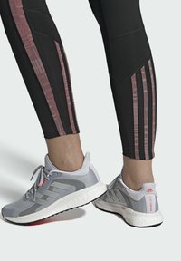 Černé sportovní legíny s růžovými vertikálními pruhy, které jsou doplněny šedými běžeckými botami s červenými akcenty a texturovými podrážkami.
