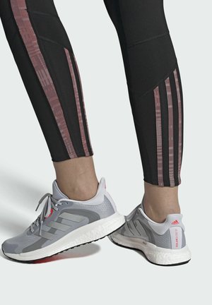 Černé sportovní legíny s růžovými vertikálními pruhy, které jsou doplněny šedými běžeckými botami s červenými akcenty a texturovými podrážkami.