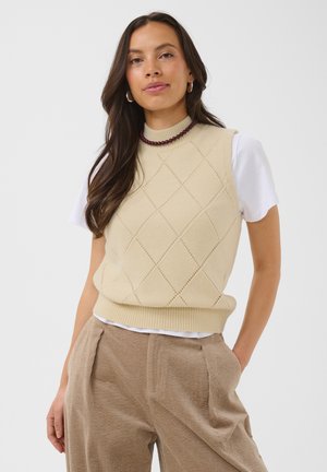 Femme aux longs cheveux bruns portant un pull sans manches beige à motif losanges sur un t-shirt blanc et un pantalon en velours côtelé beige, debout avec assurance.