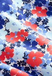 Tissu avec un motif floral comprenant des fleurs rouges, bleu foncé et bleu clair sur un fond blanc avec des plis et une texture visibles.