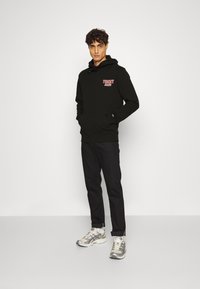 Sudadera negra con un bolsillo frontal y texto rojo "TOMMY JEANS". Combinada con pantalones negros y zapatillas grises. Tela suave, ajuste casual.