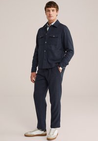 Jonge man staat in een marineblauwe jasje met knopen en klepzakken, bijpassende marineblauwe broek, witte sneakers en een wit shirt eronder.