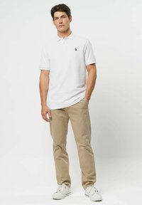 Mężczyzna stoi, ubrany w białą koszulkę polo, beżowe spodnie i białe sneakersy, z jedną ręką w kieszeni na jednolitym białym tle.