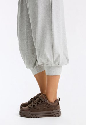 Bas de jambes portant un pantalon ample gris clair resserré aux mollets et des baskets épaisses marron foncé sur fond blanc.