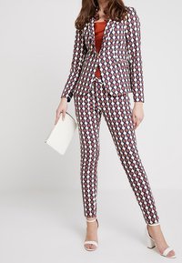 Ensemble comprenant un blazer ajusté et un pantalon slim blancs ornés de motifs circulaires multicolores. Assorti avec un haut orange et une pochette blanche.