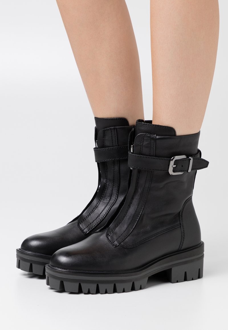 Bottines en cuir noir avec semelles crantées épaisses, couture verticale sur le devant et sangles latérales réglables avec boucles argentées sur jambes nues.