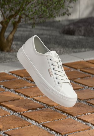 Sneaker bianco Calvin Klein con lacci sospeso sopra piastrelle quadrate di legno e piccoli ciottoli grigi all'aperto, vicino a un albero.