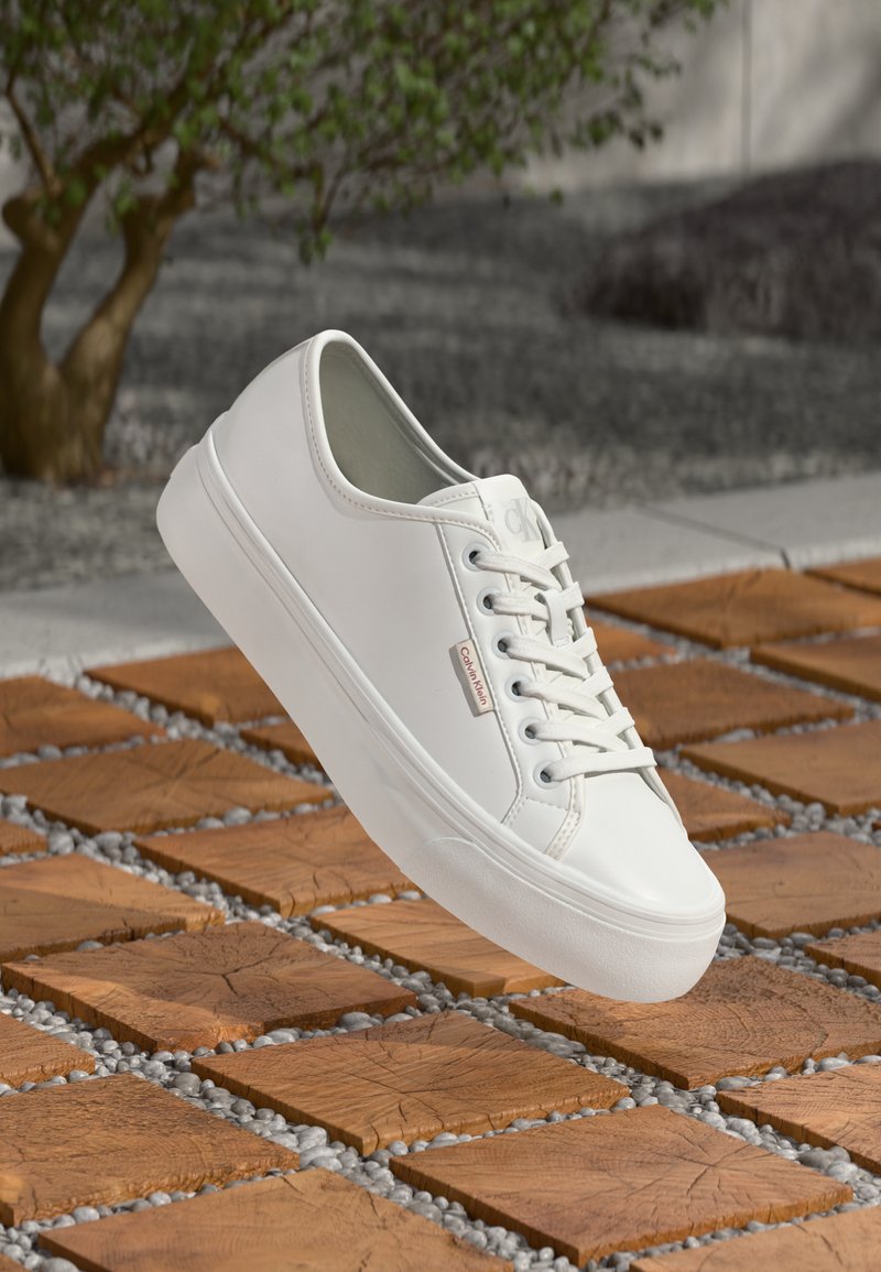 Weiße Calvin Klein Sneaker mit Schnürsenkeln, die über quadratische Holzdielen und kleine graue Steine im Freien neben einem Baum schweben.