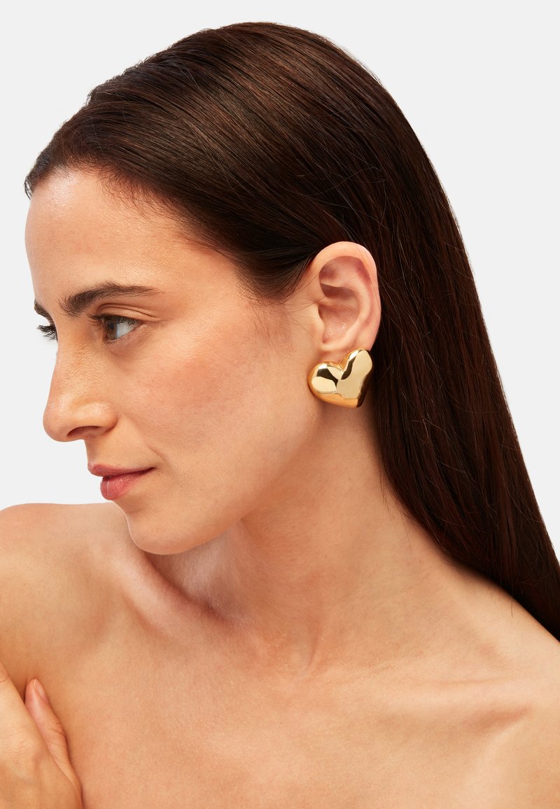 Guld hjärtformad öronkrok med en glänsande yta, placerad på en modells öra, som visar upp mjuka konturer och rundade kanter.