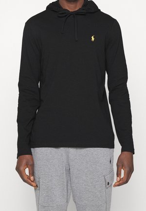 Mand iført sort langærmet hættetrøje med gul logo på brystet og lysegrå joggingbukser med sidelommer og knapklapper.