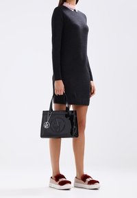 Svart lackläderhandväska med präglad logotyp, strukturerad form, topphandtag och avtagbar charm. Bärs tillsammans med en mörk klänning och fällande skor i päls.