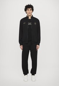 Svart zip-up jacka med guld EA7-logotyp, svart t-shirt och svarta joggers med mudd. Modellen bär vita sneakers och visar en minimalistisk stil.