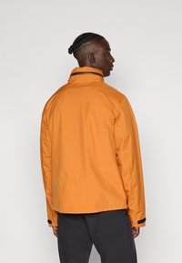 Chaqueta impermeable naranja con cuello alto, que presenta un diseño de costura diagonal y acentos de Velcro negro en los puños.