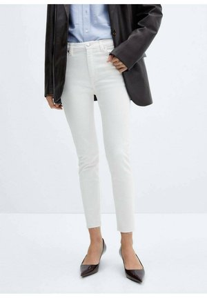 Person trägt weiße, schmal geschnittene, gekürzte Jeans, schwarze spitze Absätze, hellblaues Hemd und dunkelgrauen Blazer vor schlichtem Hintergrund.