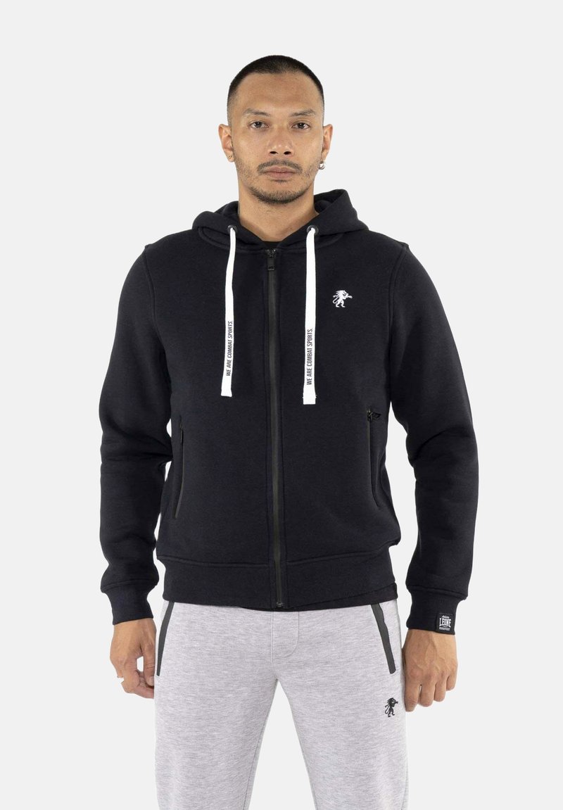 Homme portant un sweat à capuche noir zippé avec des cordons blancs et un pantalon de jogging gris clair, debout devant un fond uni clair.