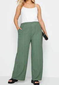 Široké kalhoty z texturované zelené tkaniny se bočními kapsami, spárované s bílým topem a černými sandály, doplněné kabelkou.