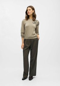 Beiger Pullover mit schwarzer Blumenstickerei an der Schulter, kombiniert mit dunkelgrauen, plissierten Hosen. Glatte Textur, lange Ärmel, lässige Passform.