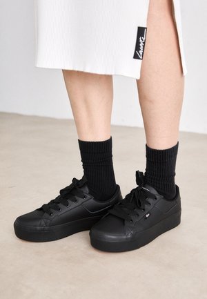 Black platform sneakers with a smooth synthetic material, rounded shape, and lace-up design. Worn with textured black crew socks.