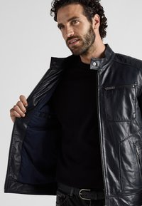 Marinblå läderjacka med dragkedjestängning, kviltade axelaccenter och en bröstficka, fodrad i marinblått tyg. Modellen håller jackan öppen.