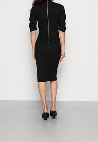 Robe noire ajustée avec un col montant, des manches trois-quarts et une fermeture éclair dorée visible courant le long du dos. Texture lisse, longueur genou.