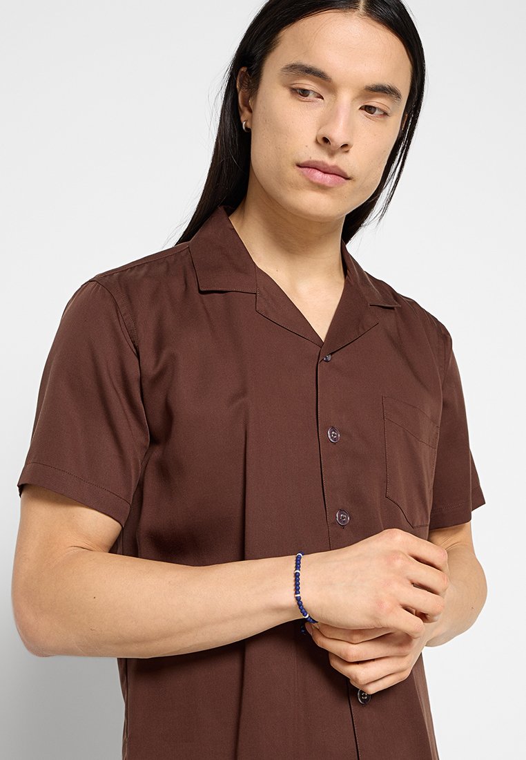 Brązowa koszula z krótkim rękawem, z kieszonką i zapięciem na guziki, wykonana z gładkiego materiału. Niebieska bransoletka z koralików na nadgarstku.