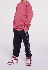 Bambino che indossa un ampio felpa rosa, pantaloni sportivi neri con strisce bianche ai lati e sneakers alte rosso e bianche, in piedi con le mani nelle tasche.