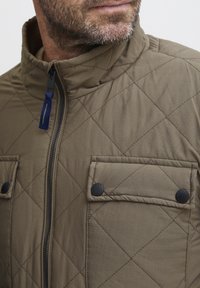 Brown quilted jacket with a high collar, zip closure, and black snap buttons on chest pockets. Features diagonal stitching and a smooth texture.