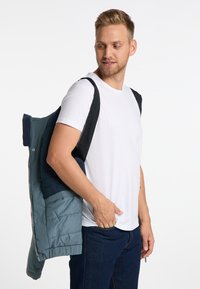 Gepolsterte Jacke in Teal und Marineblau mit einem Reißverschluss, getragen über einem weißen T-Shirt, das an einer Schulter gehalten wird. Baumwolljeans vervollständigen das Outfit.