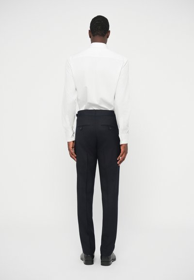 Fekete testre szabott nadrágot, fehér, hosszú ujjú inget és fekete öltönycipőt viselő férfi áll háttal egy fehér háttér előtt.