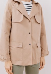 Beige vattentät jacka med en bred krage, tryckknappar och två framfickor. Strukturerad tyg med en medellång klippning och ärmsnoddar.