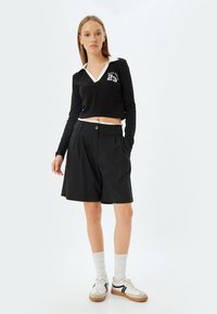 Musta lyhythihainen cropped-paita, jossa on valkoinen kaulus ja graafinen kuvio. Mustat laskostetut shortsit, joissa on napitus. Valkoiset sukat ja mustavalkoiset lenkkarit.