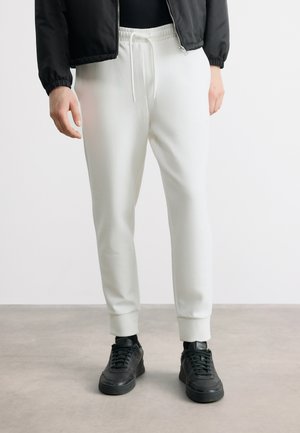 Henkilö, jolla on valkoiset kiristysnauhalliset collegehousut, mustat lenkkarit, mustat sukat ja musta vetoketjullinen takki, seisomassa harmaalla lattialla.