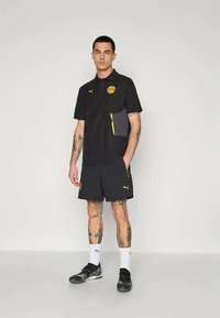 Svart polo shirt med gula accenter och logotyp, i kombination med matchande svarta shorts. Har fickor och sportig design. Bärs med svarta skor och vita strumpor.