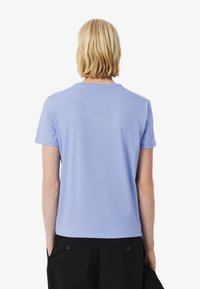 Een persoon met schouderlang blond haar draagt een lichtpaarse t-shirt, gezien van achteren tegen een witte achtergrond.