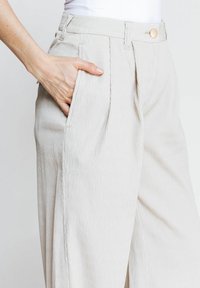 Beige, strukturierte Hosen mit weitem Bein und hoher Taille, ausgestattet mit Falten und seitlichen Taschen. Abgeschlossen mit einem Knopfverschluss an der Taille.