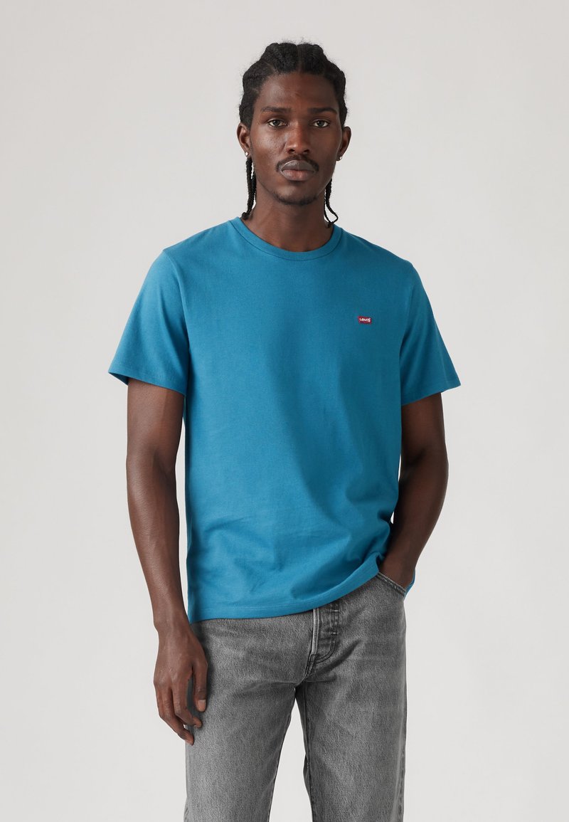 Hombre con cabello trenzado, vistiendo una camiseta azul de Levi's y vaqueros grises, de pie contra un fondo claro y liso con una mano en el bolsillo.