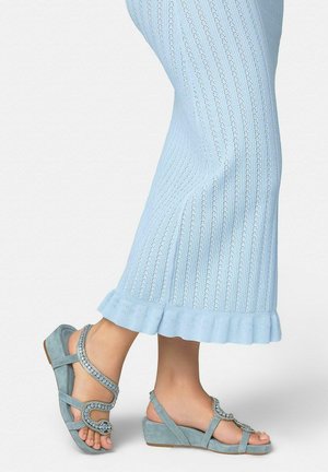 Pieds portant des sandales compensées bleu clair ornées de strass, associées à un pantalon en maille côtelée bleu clair avec des ourlets à volants.