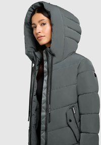 Femme portant un manteau d'hiver rembourré gris avec une grande capuche et des poches zippées, se tenant devant un fond clair.