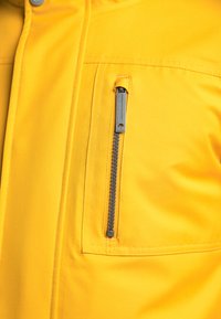 Tejido de chaqueta amarilla con una textura suave, que cuenta con un bolsillo en el pecho con cremallera, cremallera metálica y costuras en contraste.